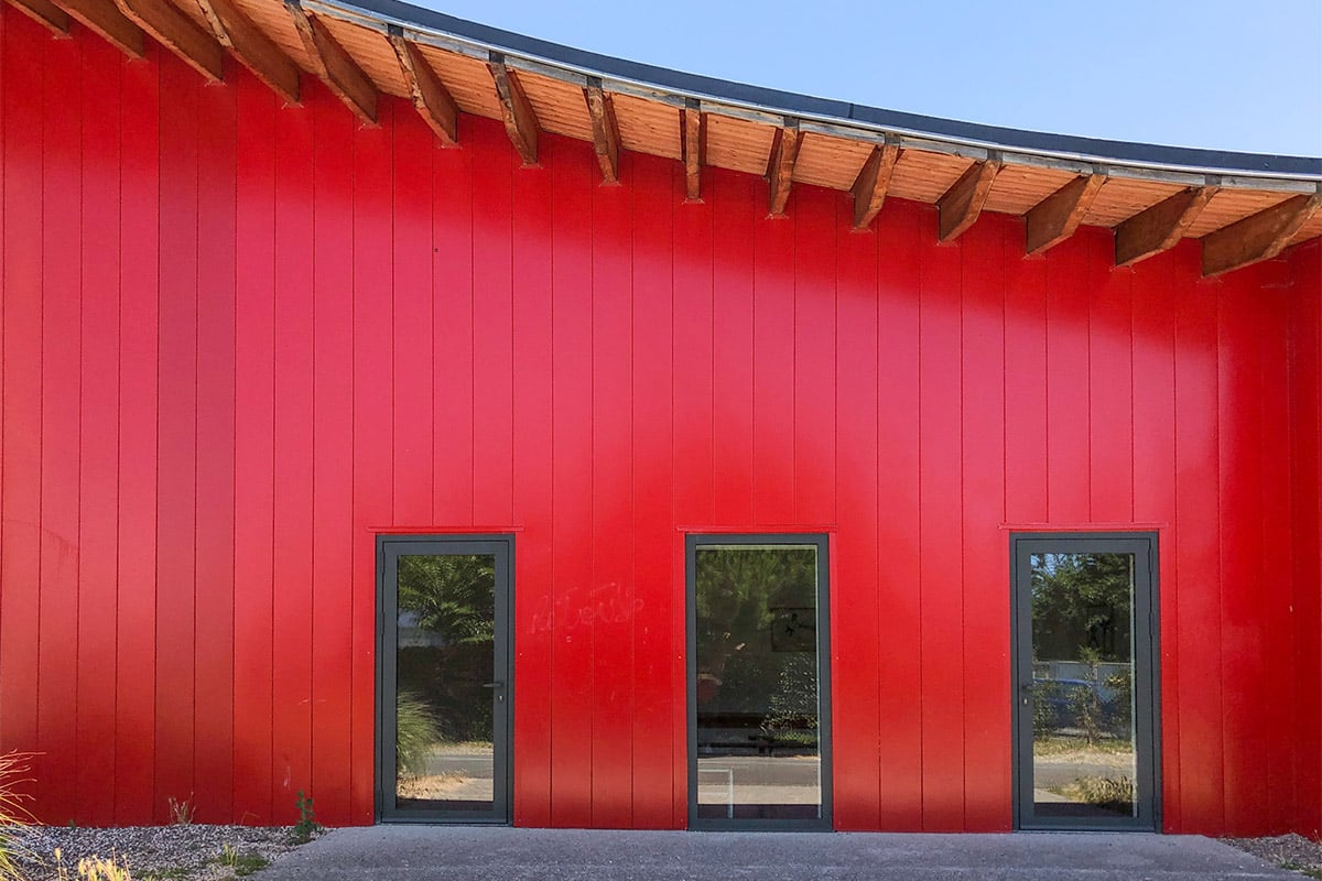 Vue de face de la façade rouge éclatante et du toit en forme de vague du dojo Pierre de Coubertin réalisé à la Teste-de-Buch par l'agence Bulle Architectes.
