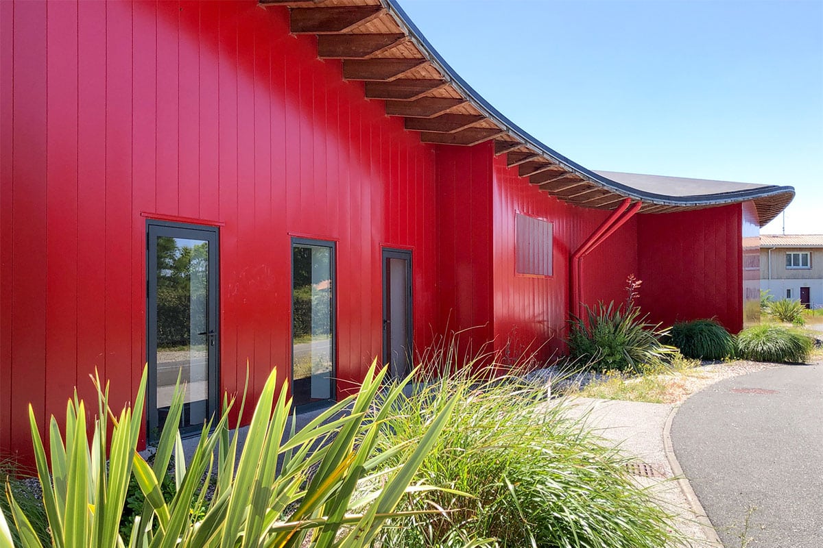 Façade rouge avec toiture en forme de vague et végétation environnante du dojo Pierre de Coubertin réalisé à la Teste-de-Buch par l'agence Bulle Architectes.