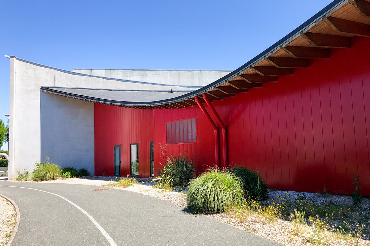 Façade rouge avec le toit en vague du dojo Pierre de Coubertin réalisé à la Teste-de-Buch par l'agence Bulle Architectes.