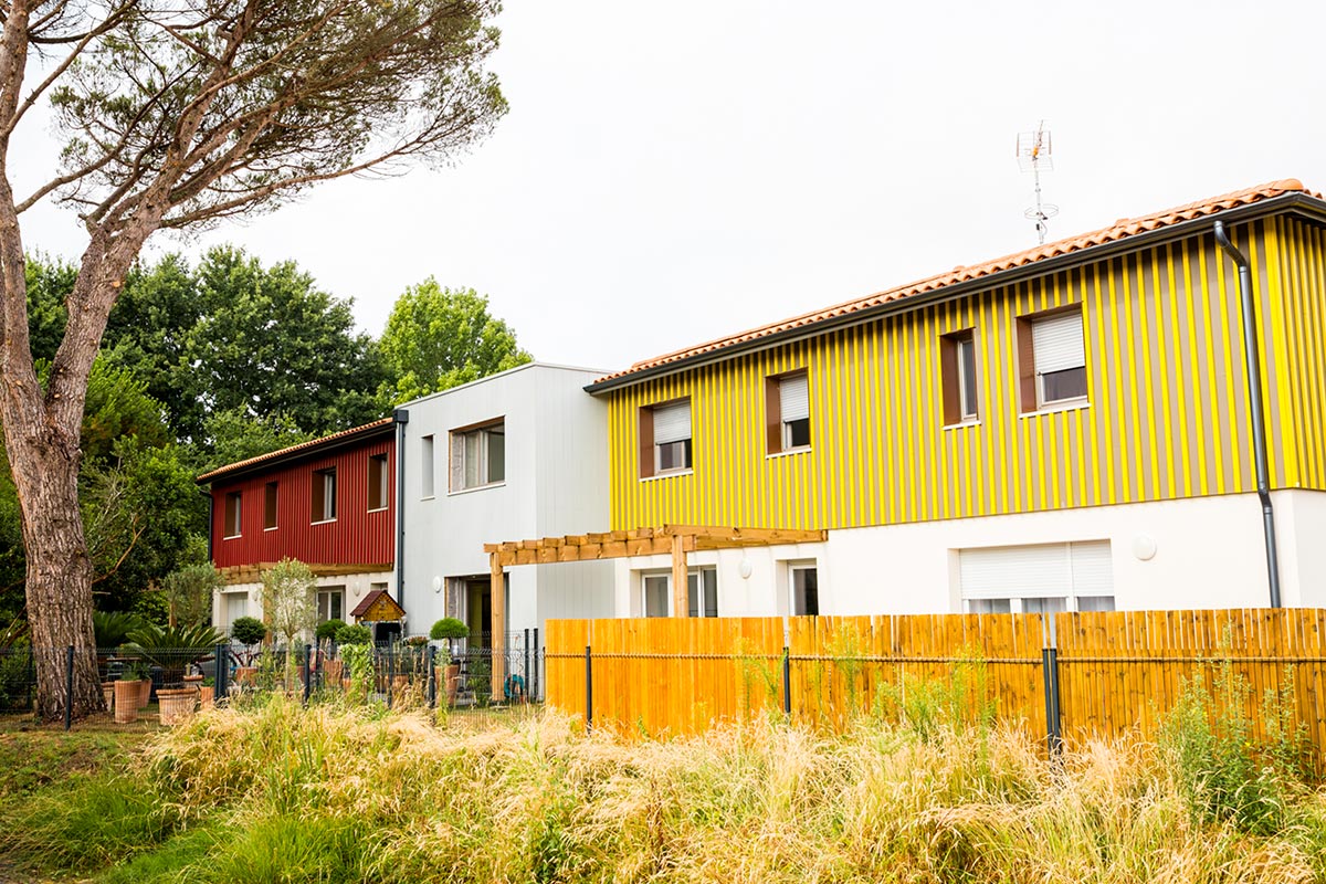 Vue sur les jardins et les façades de bâtiments au bardage en bois jaune et rouge faisant partie de la résidence L'Orée du Parc réalisée au Teich par l'agence Bulle Architectes.