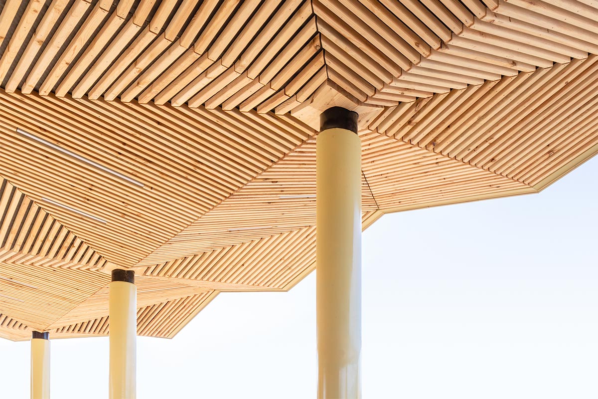 Détail des poteaux couleur doré avec les facettes en bois de la sous face de la halle réalisée au Haillan par l'agence bordelaise Bulle Architectes.